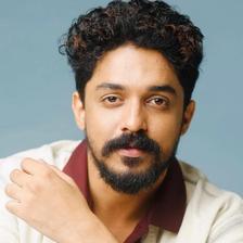 Sagar Surya with curly hair and a beard in a cream shirt over a maroon top, blue backdrop portrait