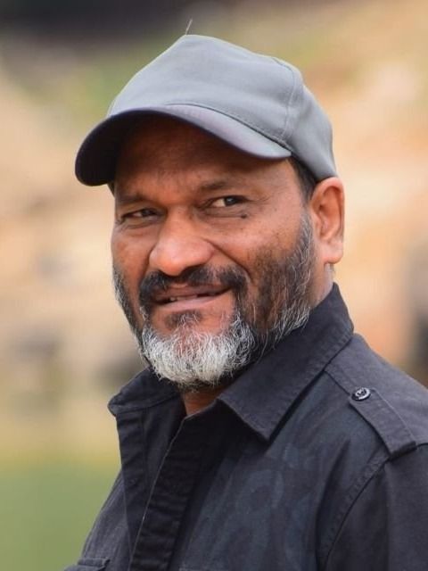 Director Sainu Chavakkadan with a grey beard, wearing a grey cap and black shirt, smiling