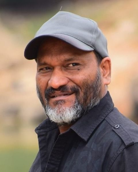 Director Sainu Chavakkadan with a grey beard, wearing a grey cap and black shirt, smiling