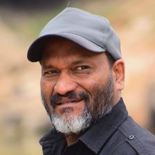 Director Sainu Chavakkadan with a grey beard, wearing a grey cap and black shirt, smiling