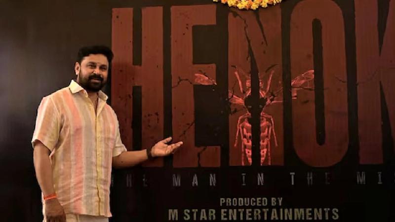 Dileep in a striped shirt points to the Henok: The Man in the Mist title board at the film's launch