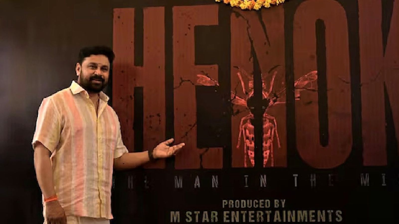 Dileep in a striped shirt points to the Henok: The Man in the Mist title board at the film's launch