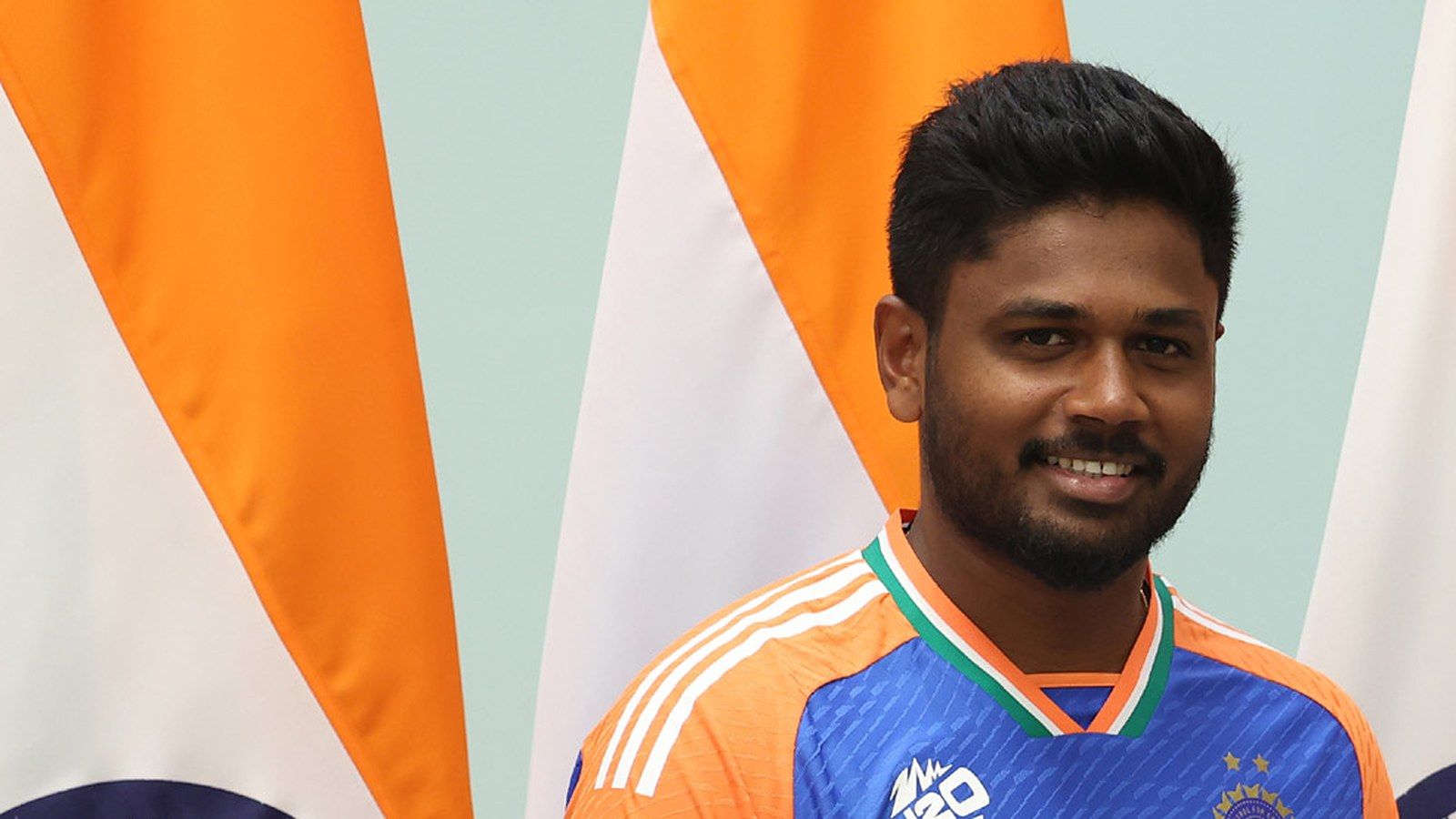 Sanju Samson smiling in the India T20 World Cup jersey in front of Indian flags.