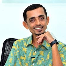 Rajesh Madhavan smiling in a printed green shirt, seated portrait