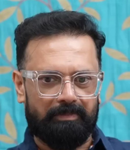 Rajeev Govindan in clear-framed glasses and a navy shirt against a teal backdrop