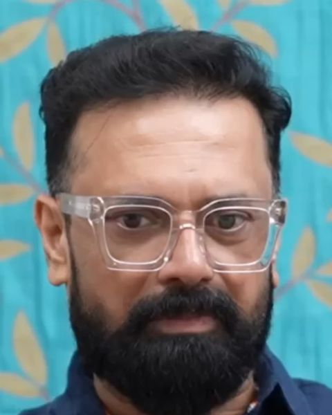 Rajeev Govindan in clear-framed glasses and a navy shirt against a teal backdrop