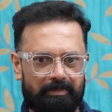 Rajeev Govindan in clear-framed glasses and a navy shirt against a teal backdrop