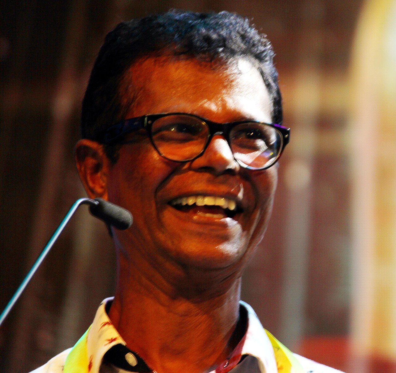 Indrans laughing in glasses beside a microphone at an event