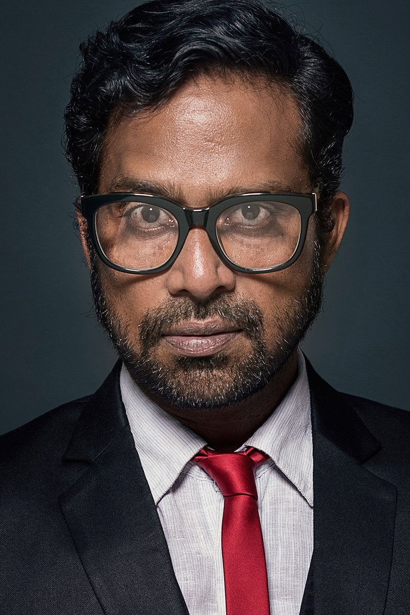 Guru Somasundaram in glasses, suit and red tie against a dark background