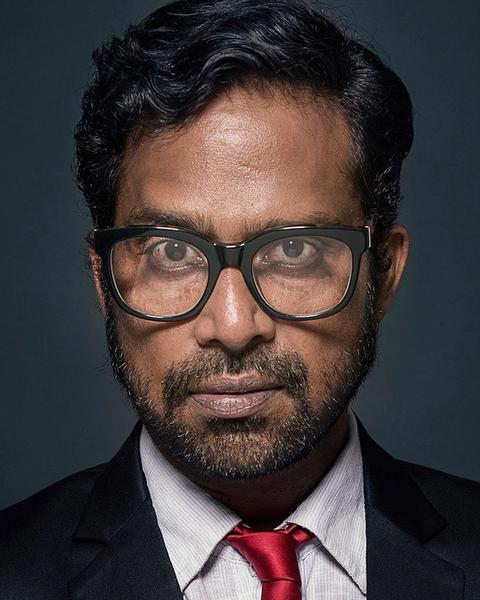 Guru Somasundaram in glasses, suit and red tie against a dark background