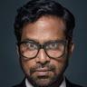 Guru Somasundaram in glasses, suit and red tie against a dark background