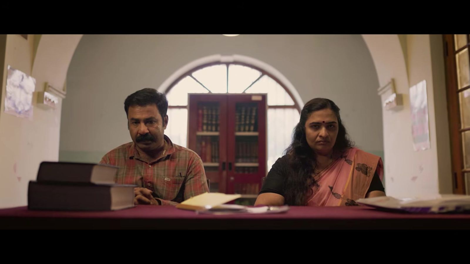 A man and a woman seated at a table in a library-like room in a frame from the Aaru Anungal trailer