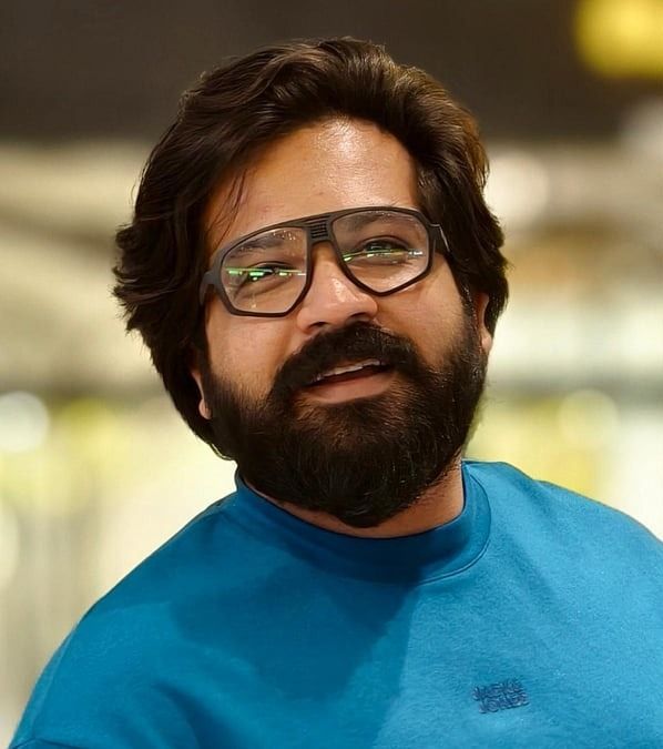 Nithish Sahadev smiling in thick-framed glasses and a teal T-shirt, with a full beard, head-and-shoulders portrait