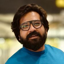 Nithish Sahadev smiling in thick-framed glasses and a teal T-shirt, with a full beard, head-and-shoulders portrait