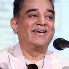 Kamal Haasan smiling at a microphone in a light jacket