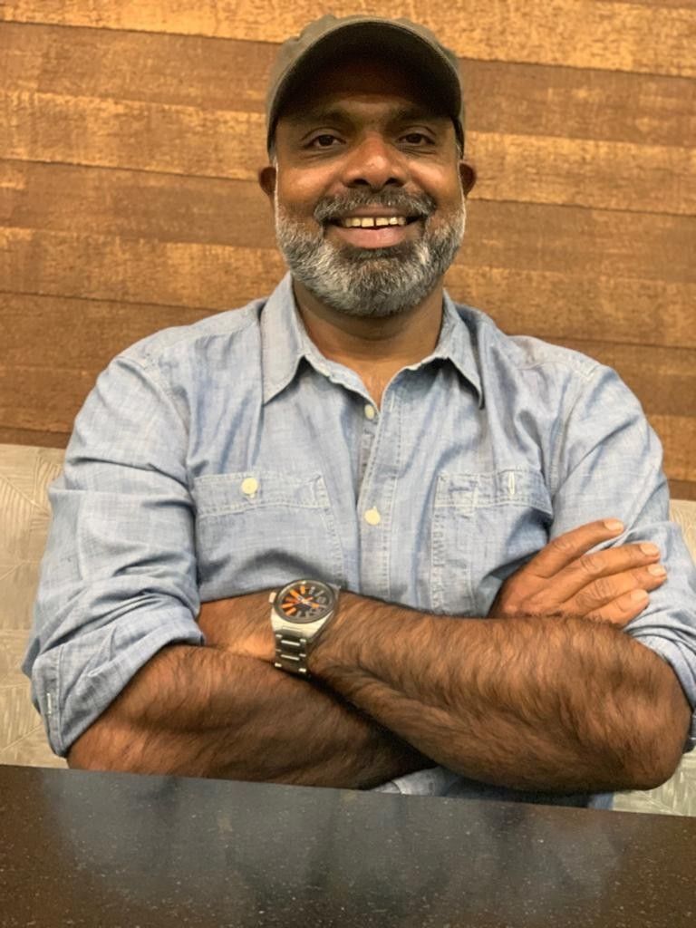 Chemban Vinod Jose smiling with a grey beard and cap, arms folded, in a light blue denim shirt and wristwatch