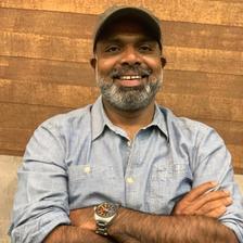 Chemban Vinod Jose smiling with a grey beard and cap, arms folded, in a light blue denim shirt and wristwatch