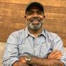 Chemban Vinod Jose smiling with a grey beard and cap, arms folded, in a light blue denim shirt and wristwatch