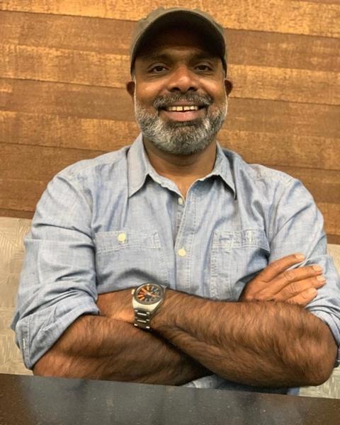 Chemban Vinod Jose smiling with a grey beard and cap, arms folded, in a light blue denim shirt and wristwatch