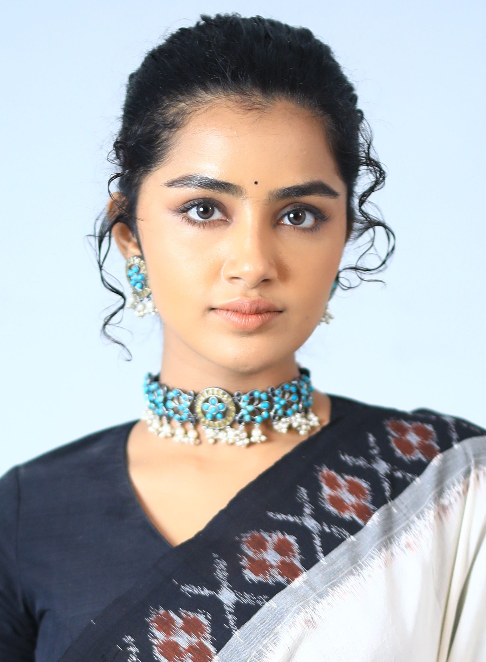 Anupama Parameswaran with her hair tied back, wearing a turquoise choker and a black-and-white ikat saree, looking at the camera against a pale blue backdrop