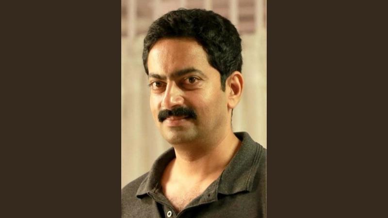 Saiju Kurup in a dark grey polo shirt, smiling slightly, on a plain dark brown background