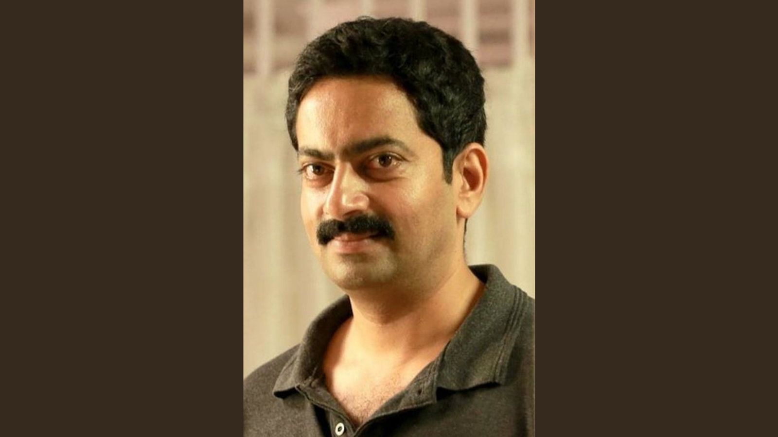 Saiju Kurup in a dark grey polo shirt, smiling slightly, on a plain dark brown background