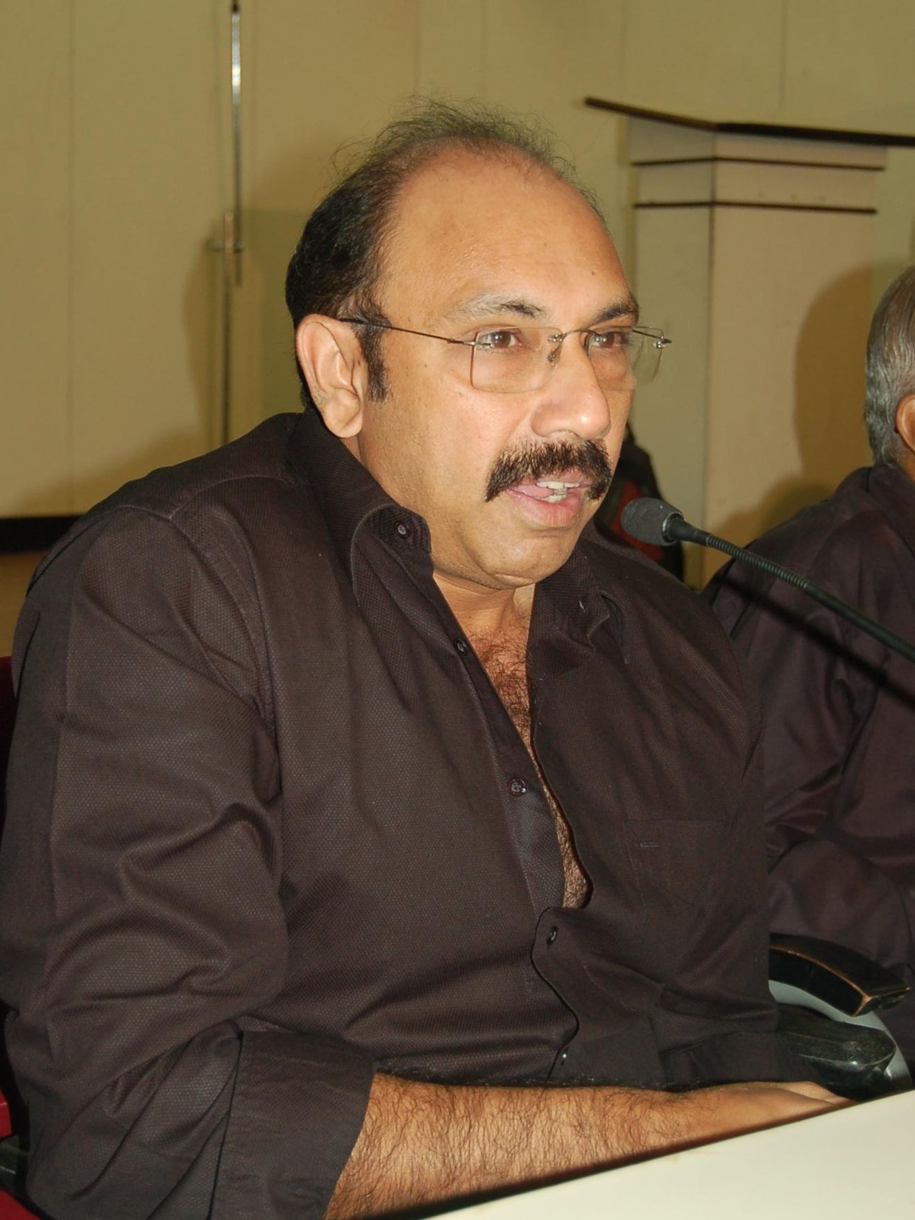 Sathyaraj in glasses speaking at a press event table