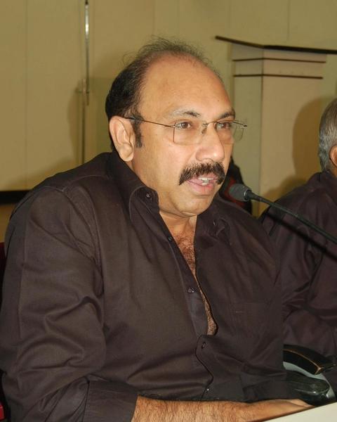 Sathyaraj in glasses speaking at a press event table