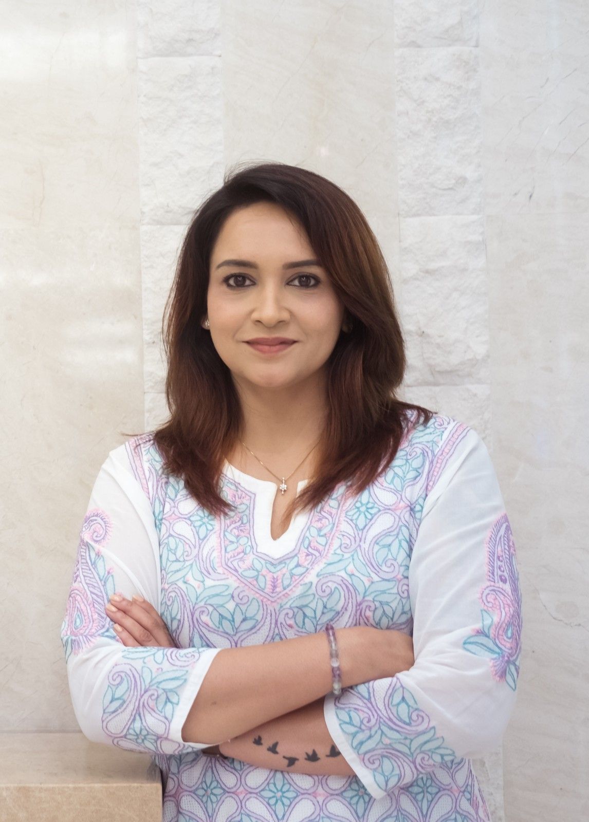 Lena smiling with arms folded, wearing a white kurta with pink and blue embroidery against a cream stone wall