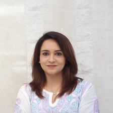 Lena smiling with arms folded, wearing a white kurta with pink and blue embroidery against a cream stone wall