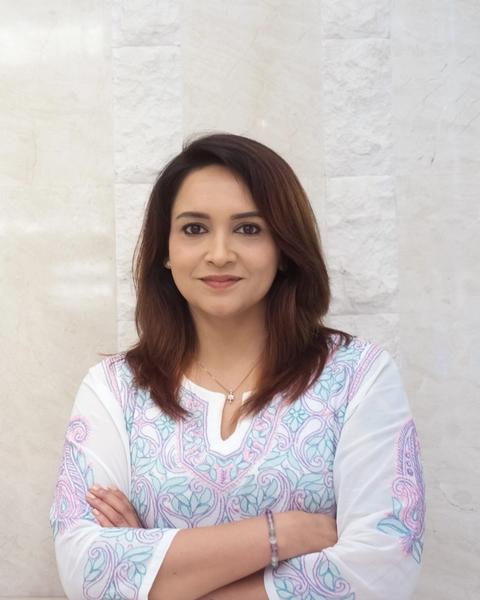 Lena smiling with arms folded, wearing a white kurta with pink and blue embroidery against a cream stone wall