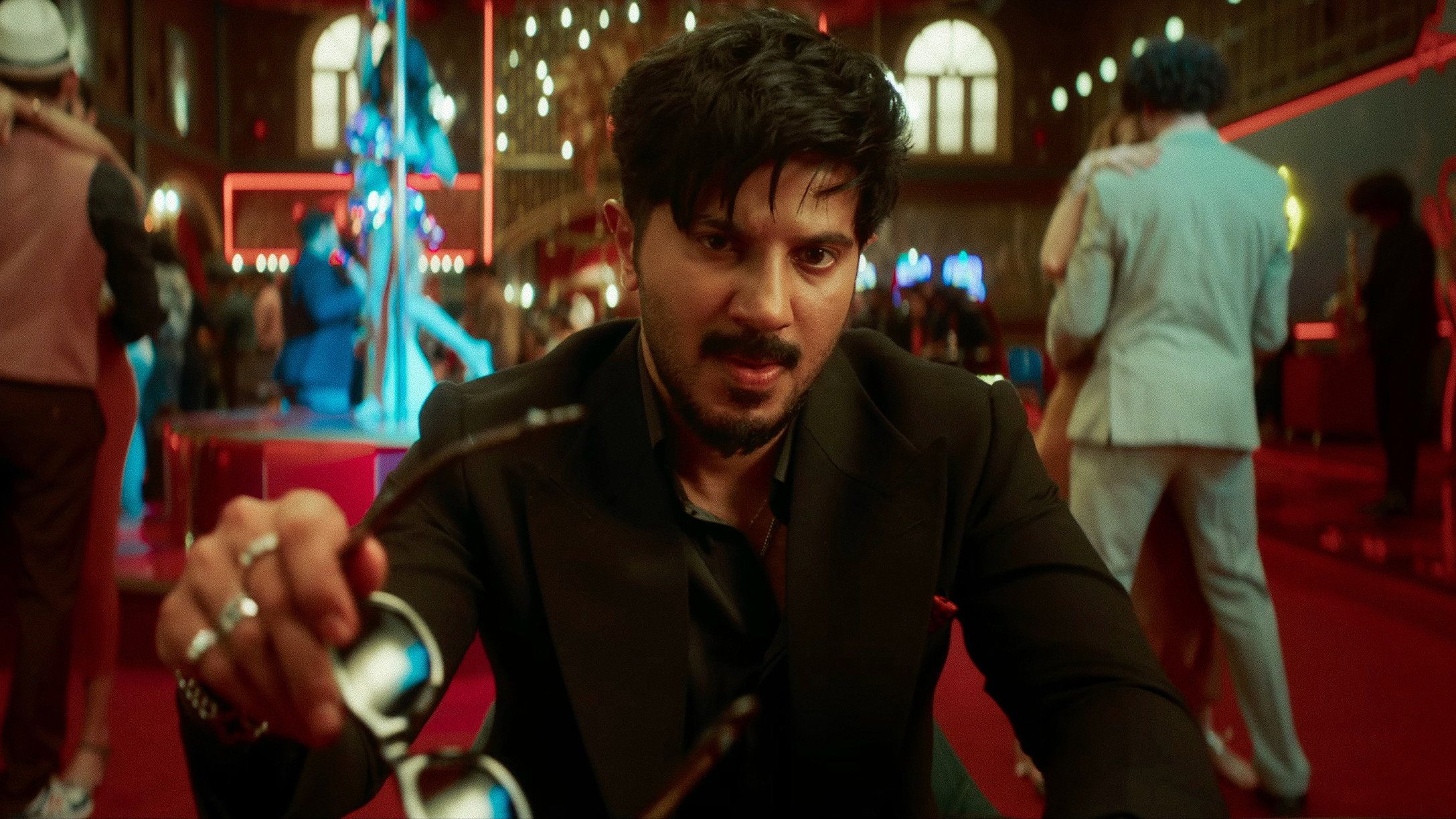 Dulquer Salmaan as Dan John holding out a pair of sunglasses in a casino in I'm Game