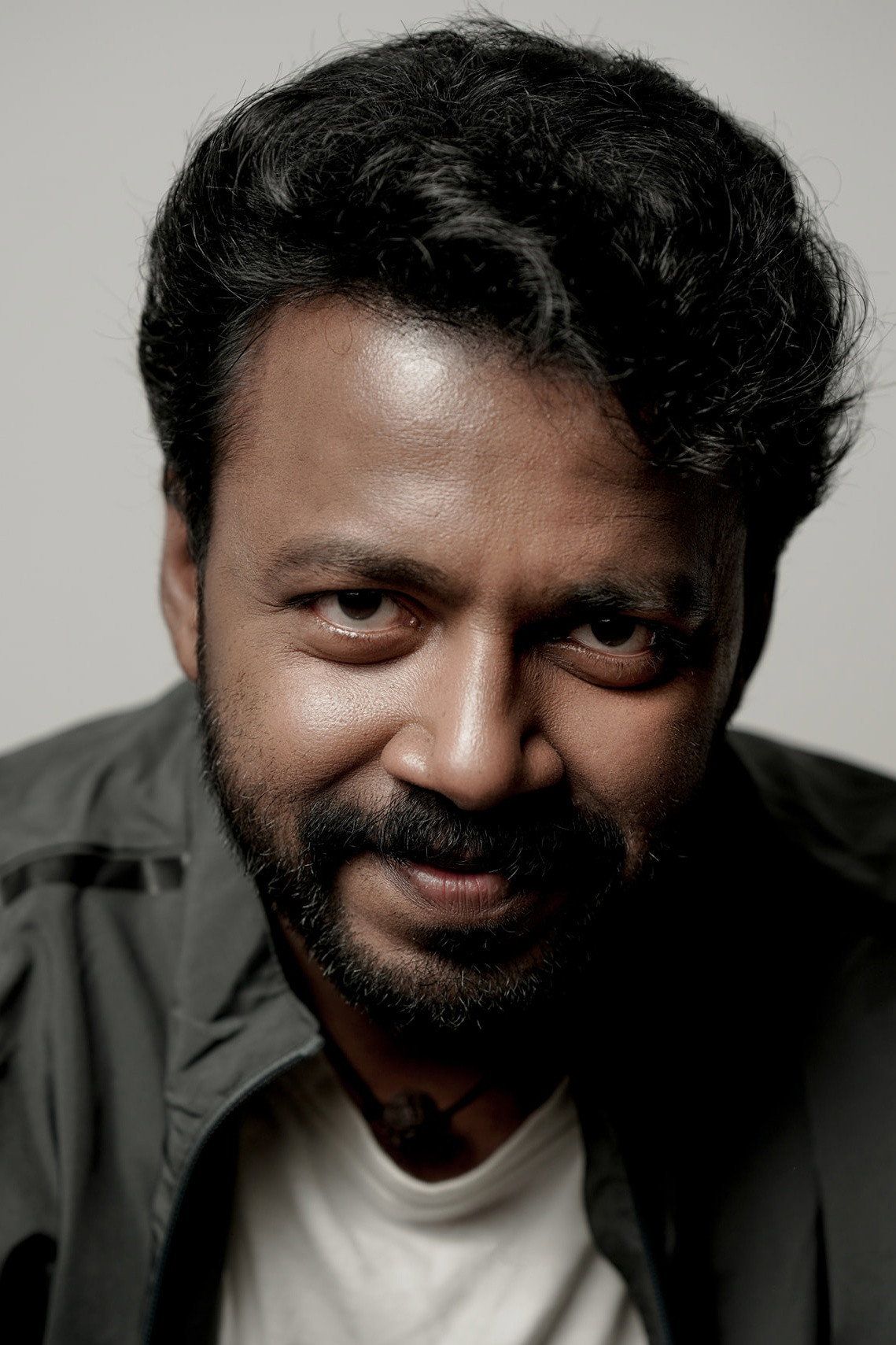 Vishnu Unnikrishnan with a beard in a dark jacket over a white T-shirt, grey backdrop portrait
