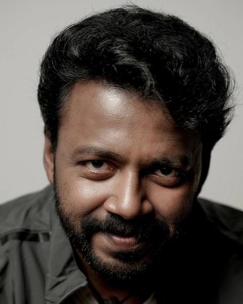 Vishnu Unnikrishnan with a beard in a dark jacket over a white T-shirt, grey backdrop portrait