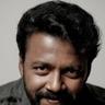 Vishnu Unnikrishnan with a beard in a dark jacket over a white T-shirt, grey backdrop portrait