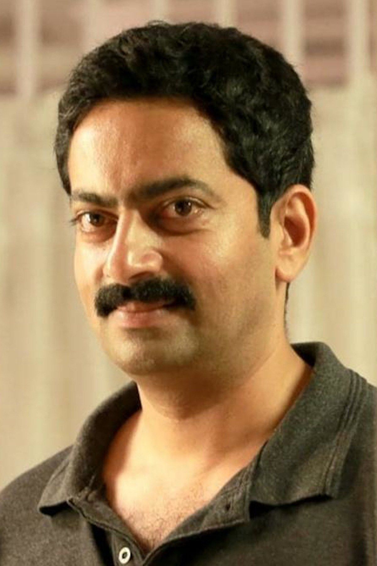 Saiju Kurup in a dark grey polo shirt, smiling slightly at the camera against a soft cream background