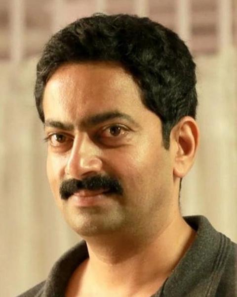 Saiju Kurup in a dark grey polo shirt, smiling slightly at the camera against a soft cream background
