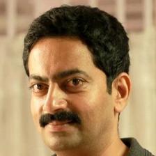 Saiju Kurup in a dark grey polo shirt, smiling slightly at the camera against a soft cream background