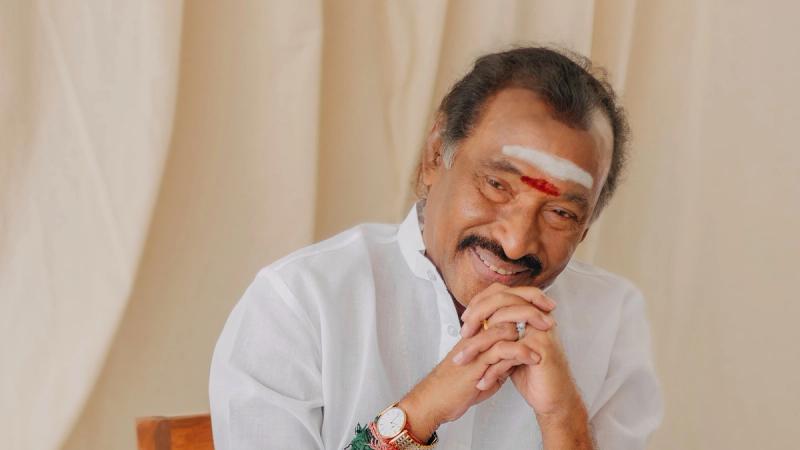 Composer Deva smiling with his hands clasped, seated in a white shirt