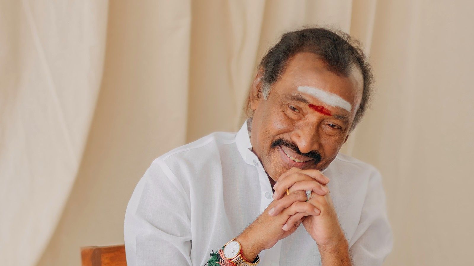 Composer Deva smiling with his hands clasped, seated in a white shirt