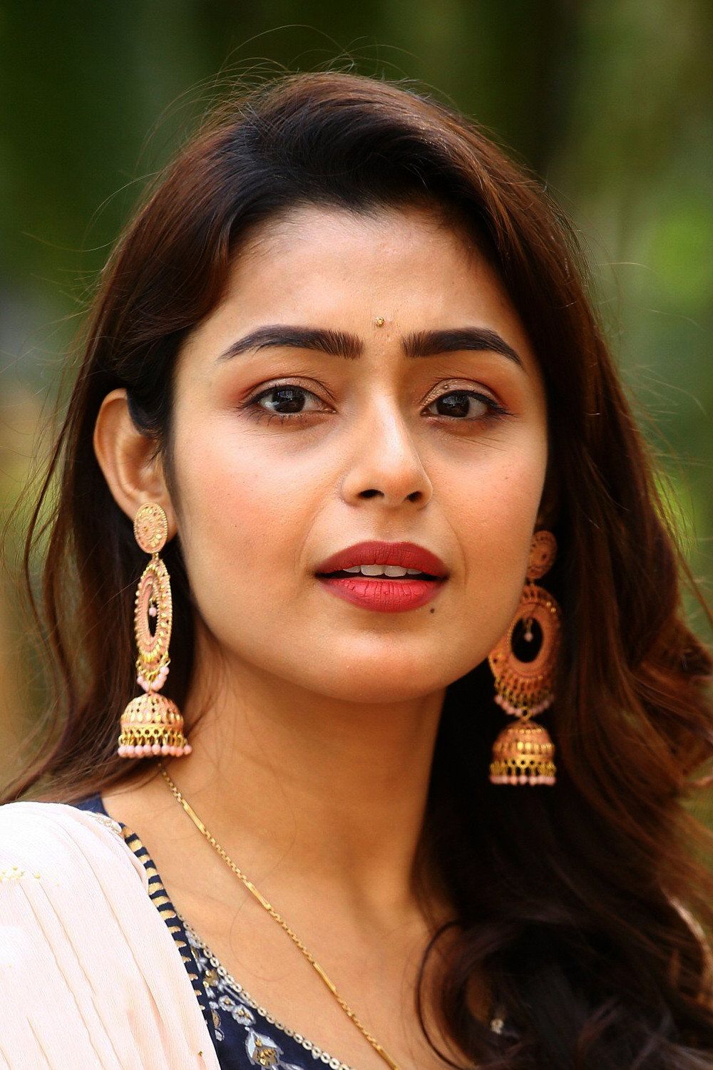 Mokksha in gold jhumkas and a white top against green foliage, portrait
