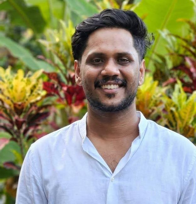 Belraj Kalarickal smiling in a white shirt in front of green and red garden plants