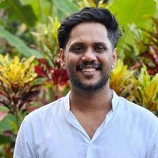 Belraj Kalarickal smiling in a white shirt in front of green and red garden plants