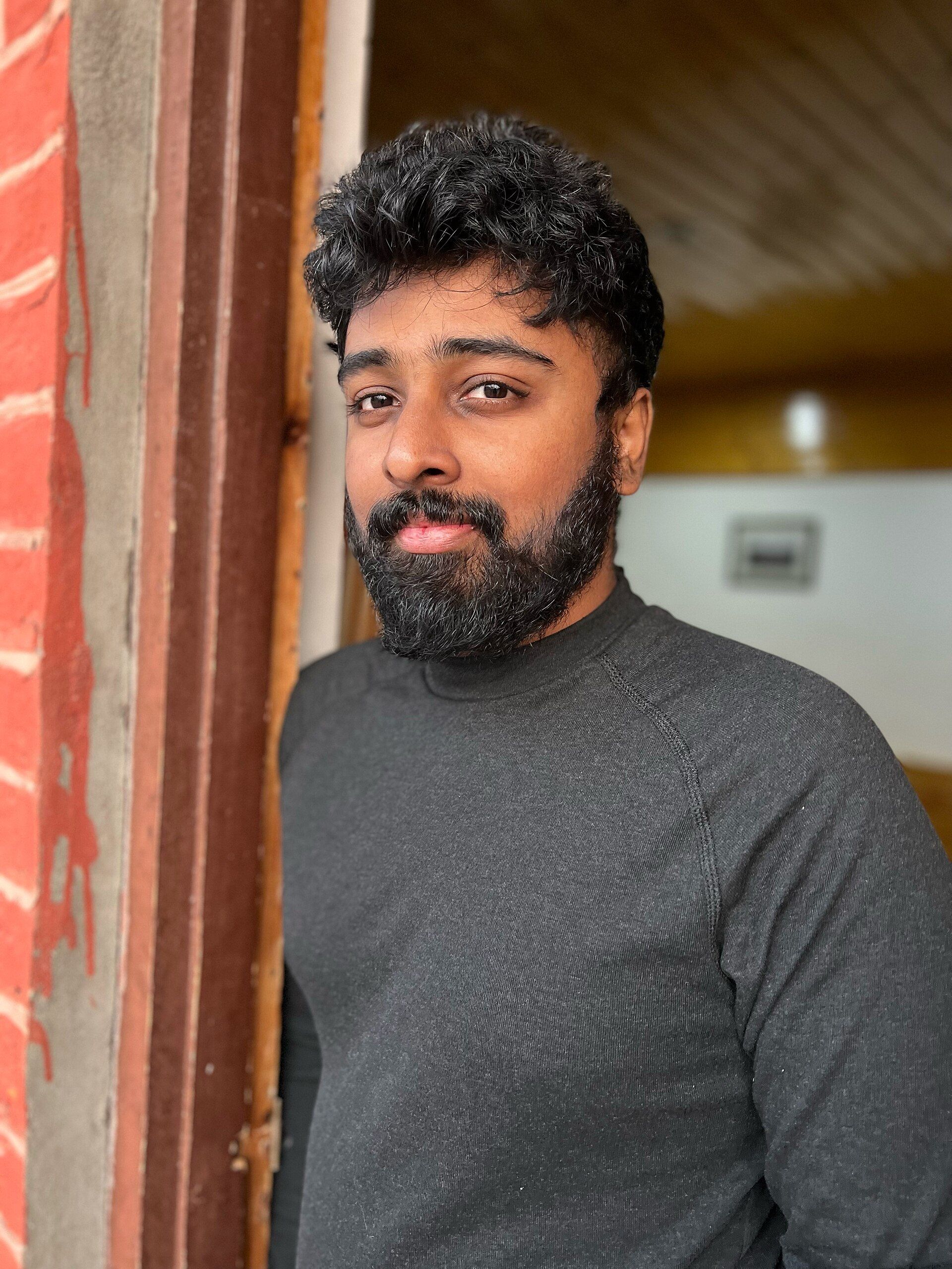 Nimish Ravi in a grey sweater leaning against a red door frame