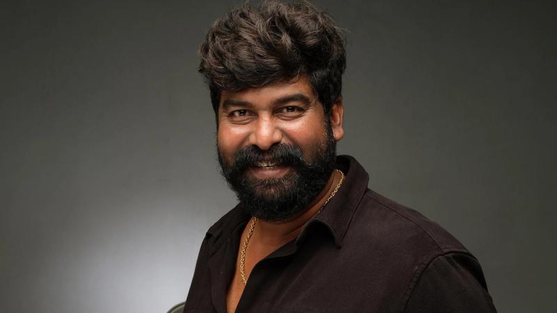 Joju George smiling in a dark shirt against a grey backdrop