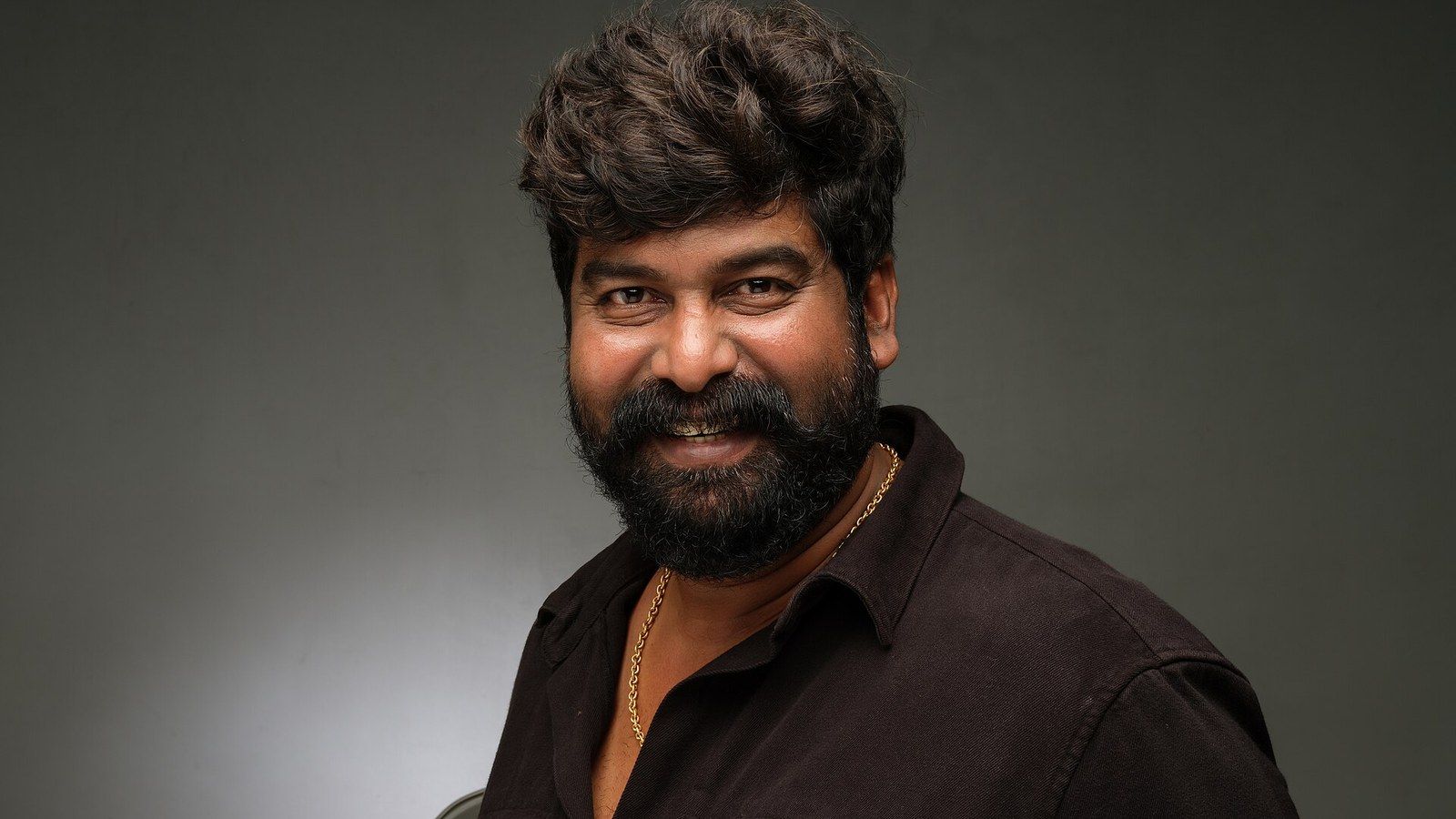 Joju George smiling in a dark shirt against a grey backdrop