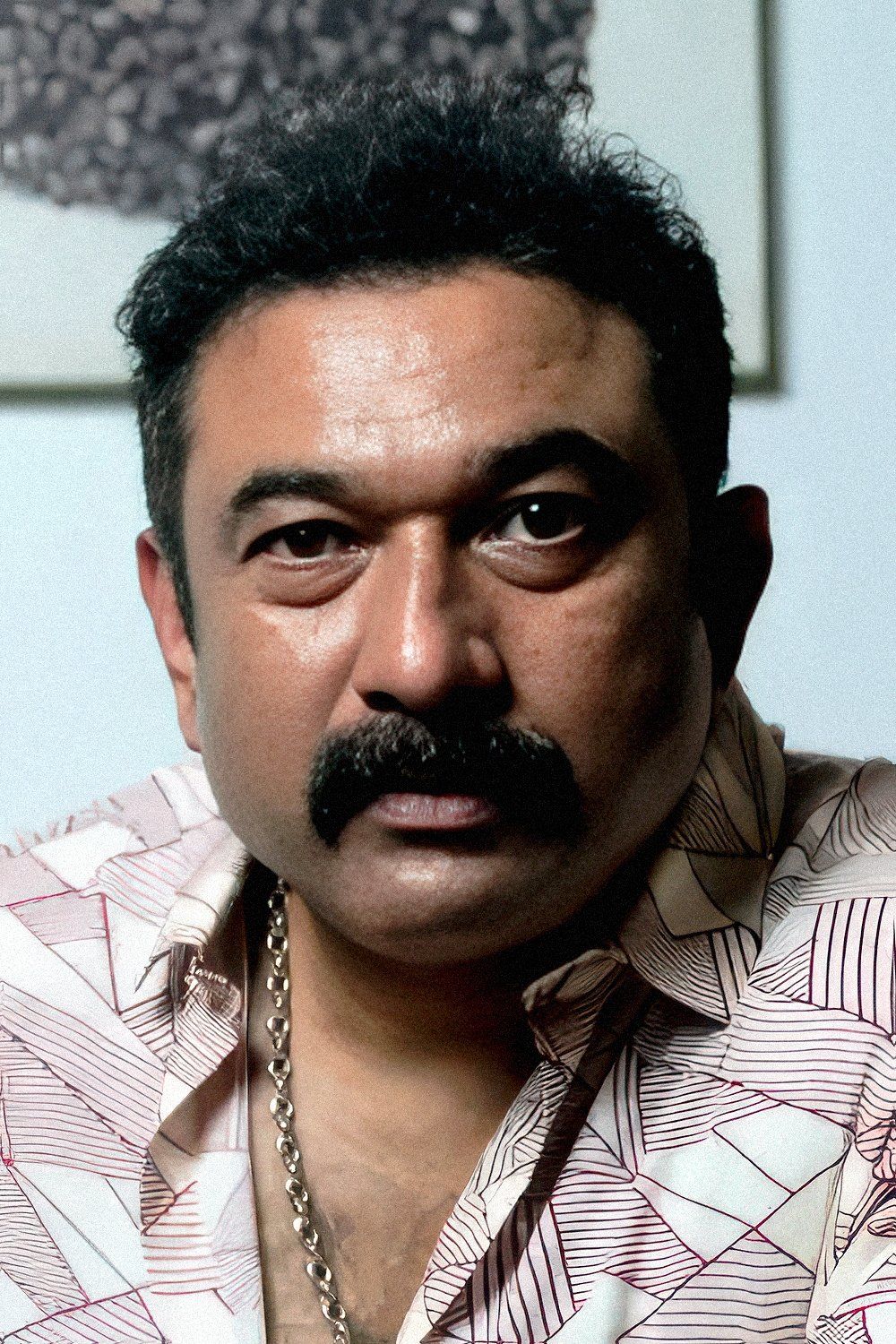Baburaj with a thick moustache, in a printed white shirt and a silver chain, looking at the camera