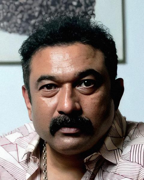 Baburaj with a thick moustache, in a printed white shirt and a silver chain, looking at the camera