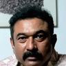 Baburaj with a thick moustache, in a printed white shirt and a silver chain, looking at the camera