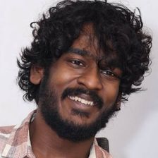 Aegan smiling with curly hair and a beard in a pink checked shirt, head-and-shoulders portrait